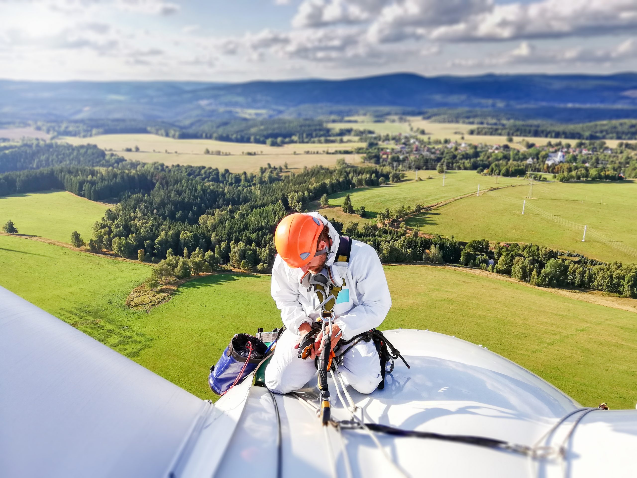 Industrial climber, rope access technician kneeling on enormous onshore wind-turbine while preparing for blade inspection with amazing view on Ore mountain (Krusne hory) moutain range